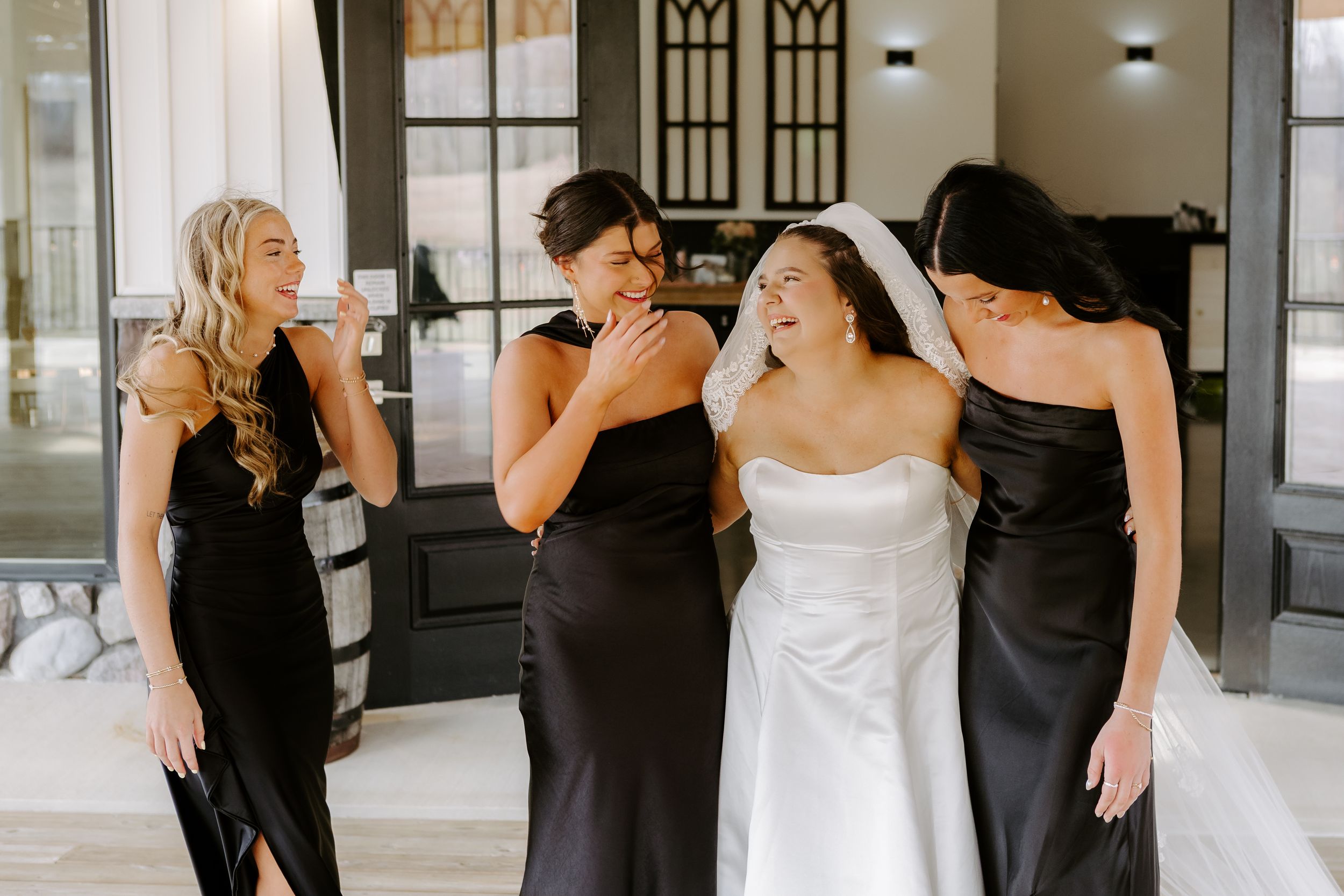 Bride in white gown laughing with three bridesmaids in black satin dresses during wedding preparations indoors.