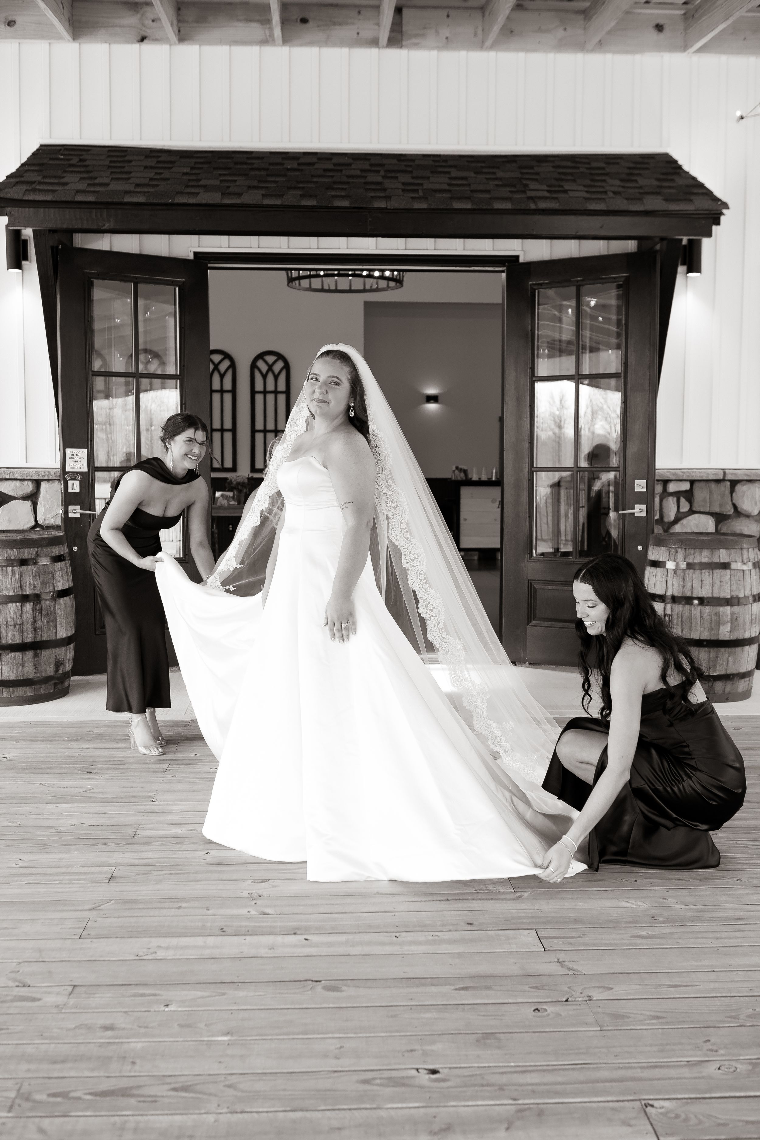 Bridesmaids helping bride adjust her wedding gown and veil on wooden deck before ceremony.