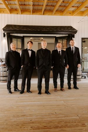 Five groomsmen in black suits pose together inside a rustic barn wedding venue with wooden floors.
