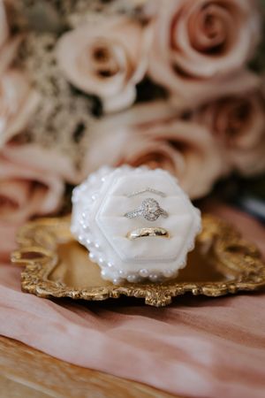 Wedding rings displayed in a white velvet box on a gold tray with blush roses in the background.