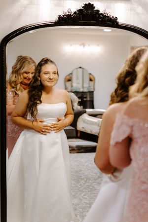 Bride in white strapless gown admires her reflection in ornate mirror while bridesmaid adjusts her dress.