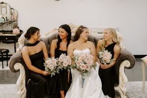 Bride in white strapless gown sits with three bridesmaids in black dresses holding pink bouquets on elegant sofa.