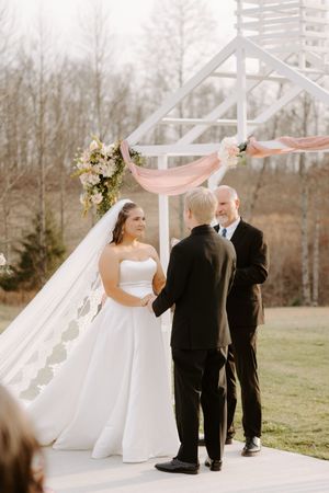 Bride and groom exchange vows under a white floral arch with pink draping at an outdoor wedding ceremony.