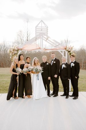 Wedding party poses in black attire with bride in white gown under a rustic white arch with floral arrangements.
