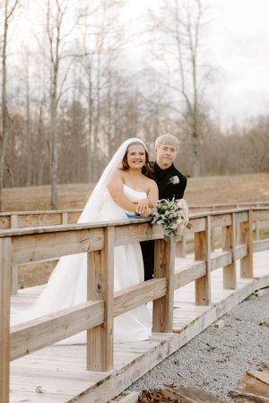 Bride and groom pose together on a rustic wooden bridge, smiling with a bouquet, surrounded by winter trees.
