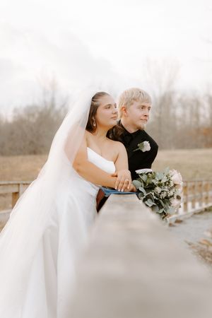 Bride in white gown and veil stands with groom in black suit on bridge holding pink bouquet outdoors.