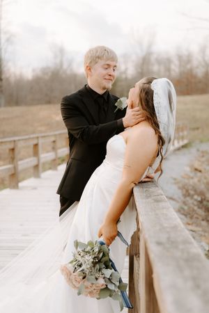Groom in black suit tenderly holds bride's face on wooden bridge, bride holds pink bouquet.