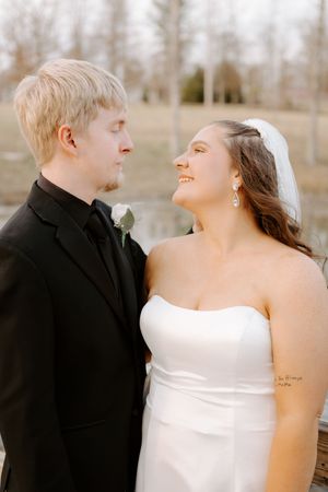A smiling bride in a white strapless gown and veil gazes at her groom in a black suit during their wedding day.