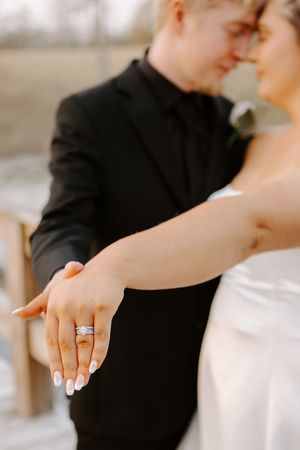 Close-up of bride showing diamond wedding ring set while groom in black suit embraces her from behind.