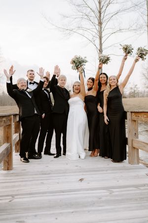 Wedding party celebrating on a wooden bridge, bridesmaids in black gowns and groomsmen in tuxedos.