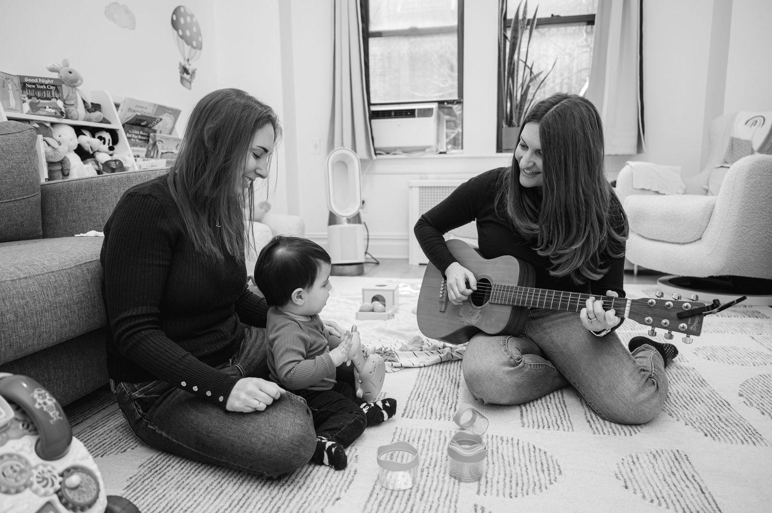 Two women and a baby sit on a play mat as one strums an acoustic guitar in a cozy living room.