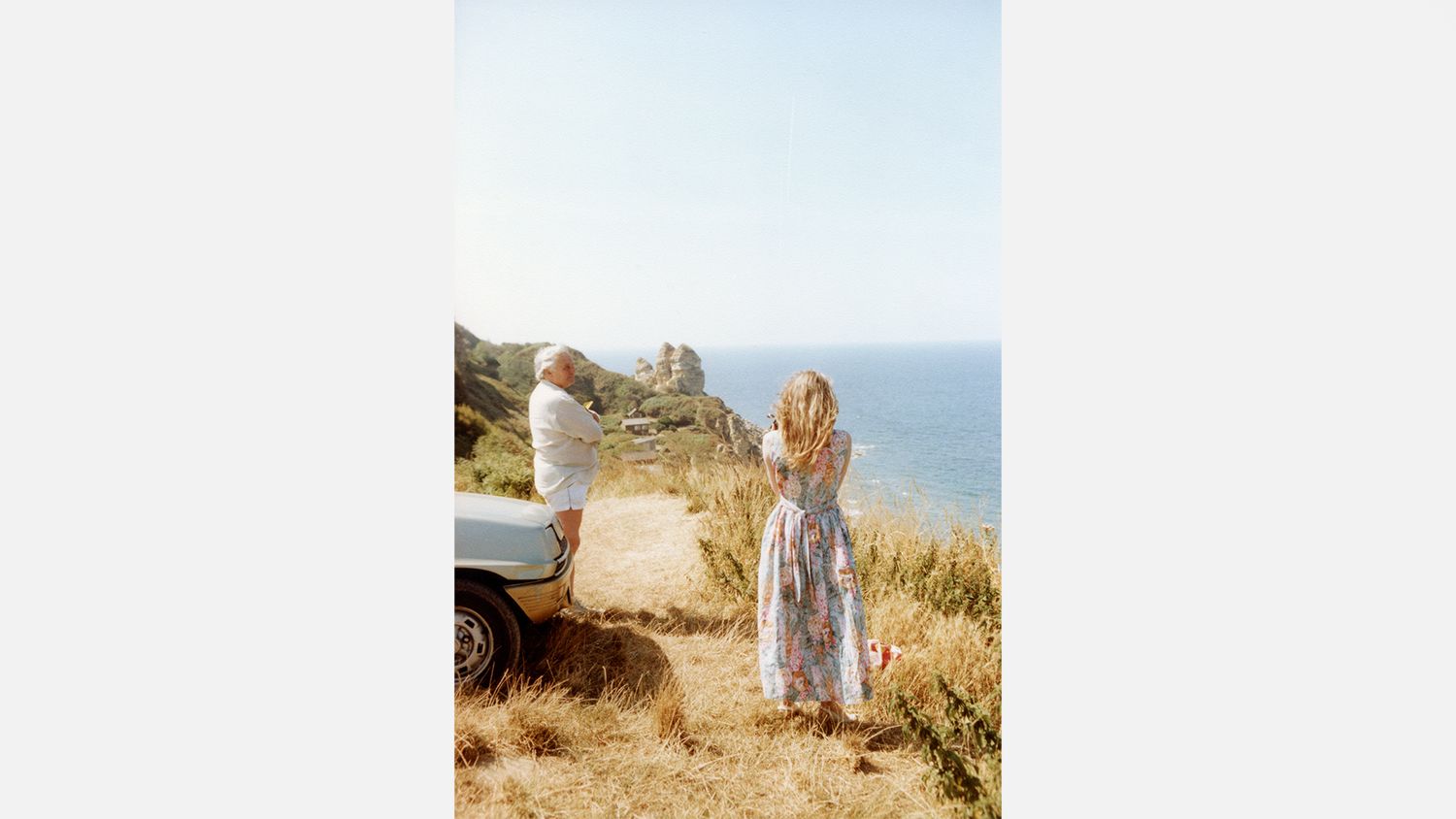 Father and daughter standing on a scenic coastal cliff beside a car, overlooking the blue sea on a sunny day.