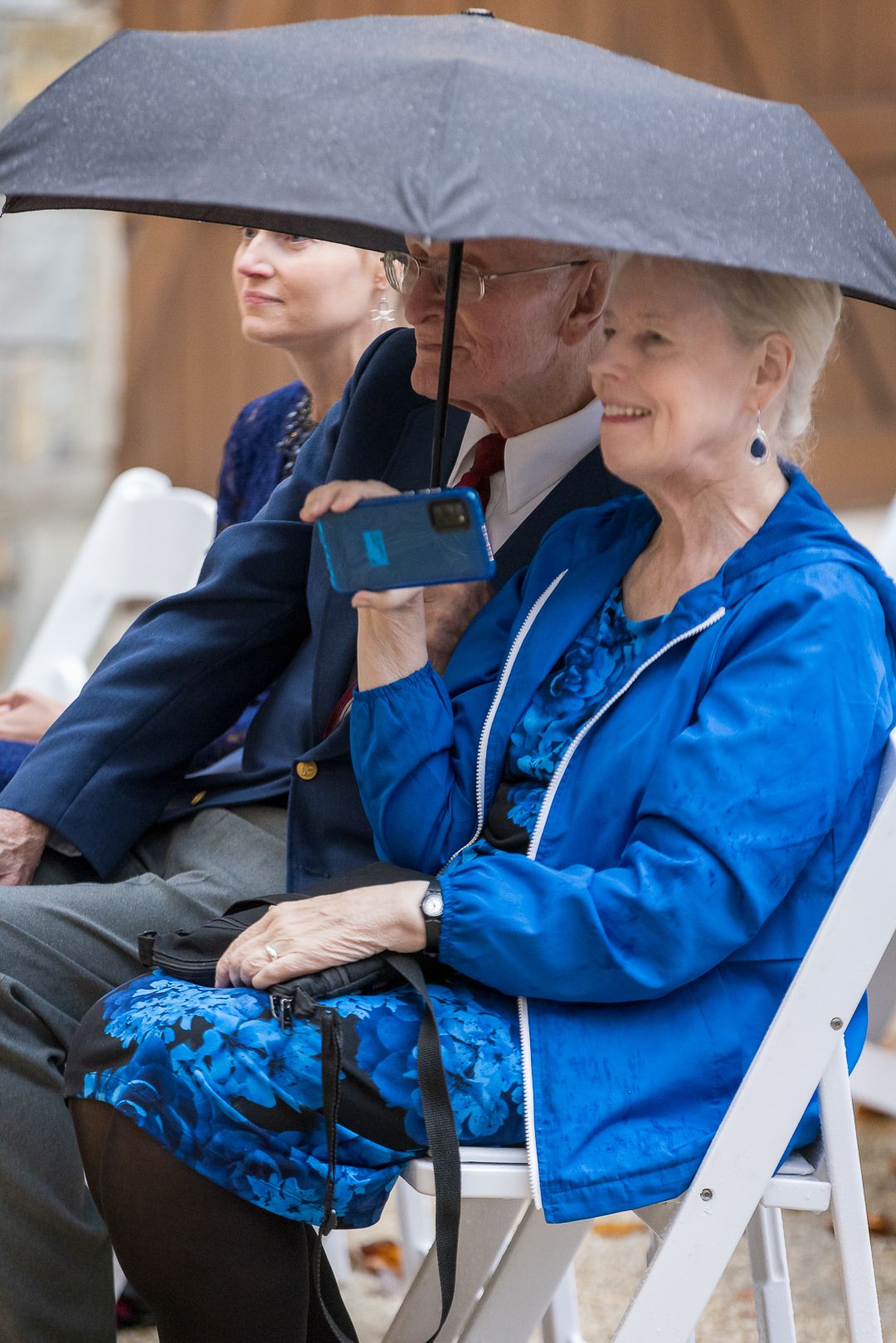Elderly couple sharing umbrella in rain at outdoor event, woman in blue jacket smiling and holding phone.