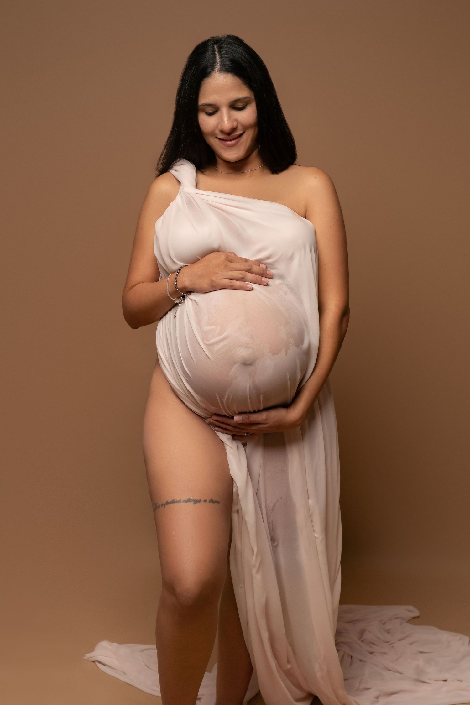 Pregnant woman in sheer white draped fabric cradling her baby bump, smiling softly against a warm beige background.
