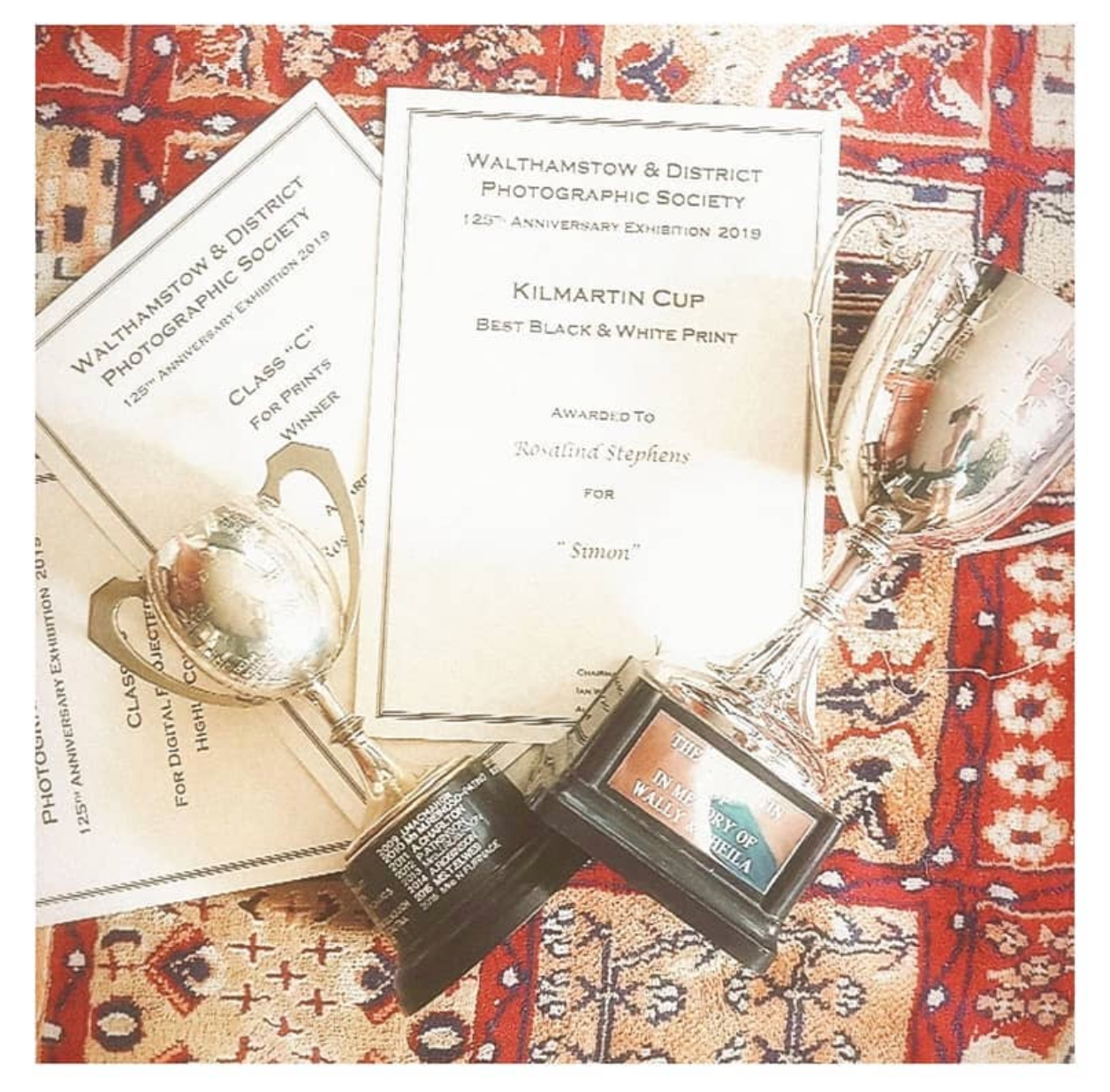Silver trophies and Kilmartin Cup award certificates from Walthamstow & District Photographic Society on a red patterned rug.