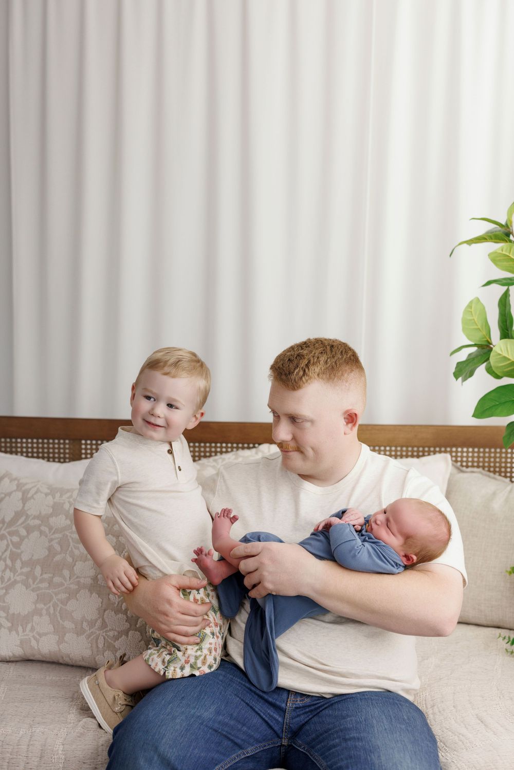 A young father sits on a couch holding a newborn baby while a smiling toddler stands beside him indoors.