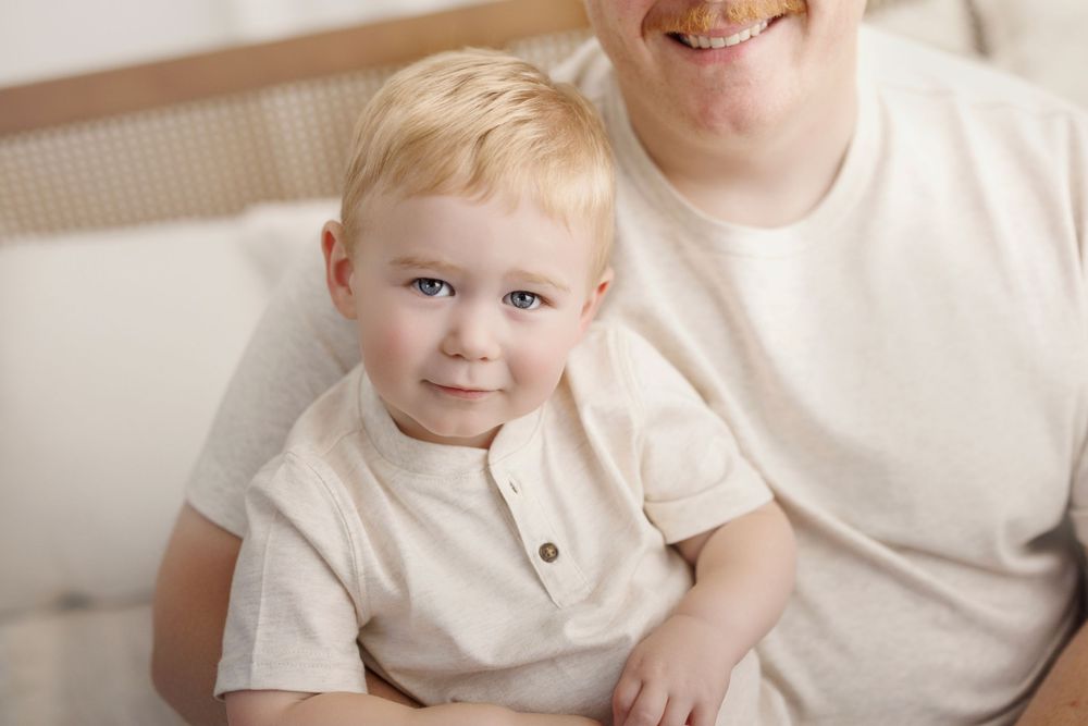 Adorable blonde toddler boy sitting on lap of adult in white shirt, smiling at camera in bright cozy setting.