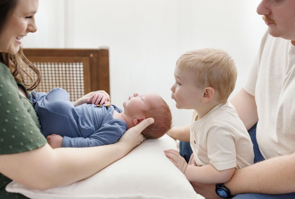 A mother holds her newborn baby while a toddler sibling and father look on during a family portrait session.