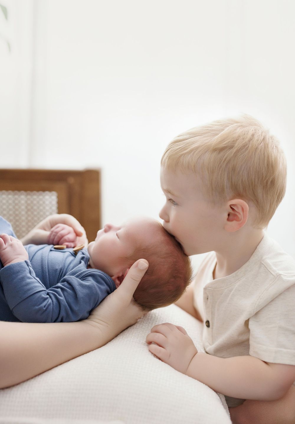 Toddler boy with blonde hair gently kissing a newborn baby in a blue onesie held by an adult.