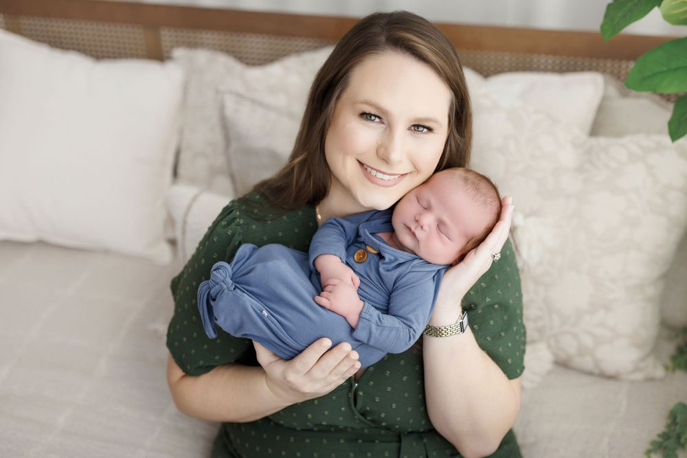 Smiling mother in green dress holds sleeping newborn wrapped in blue swaddle during professional photo session.