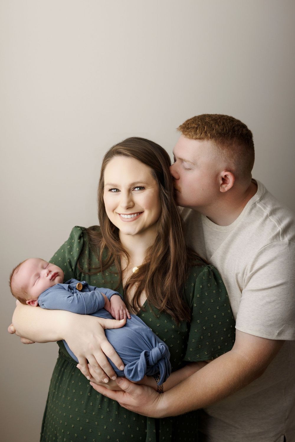 A smiling woman in a green dress holds a newborn baby while a red-haired man kisses her cheek.