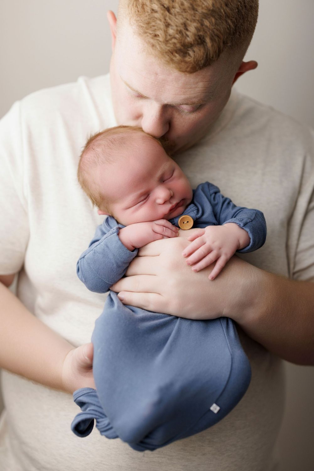 A tender moment as a red-haired father kisses his sleeping newborn baby dressed in a blue romper.