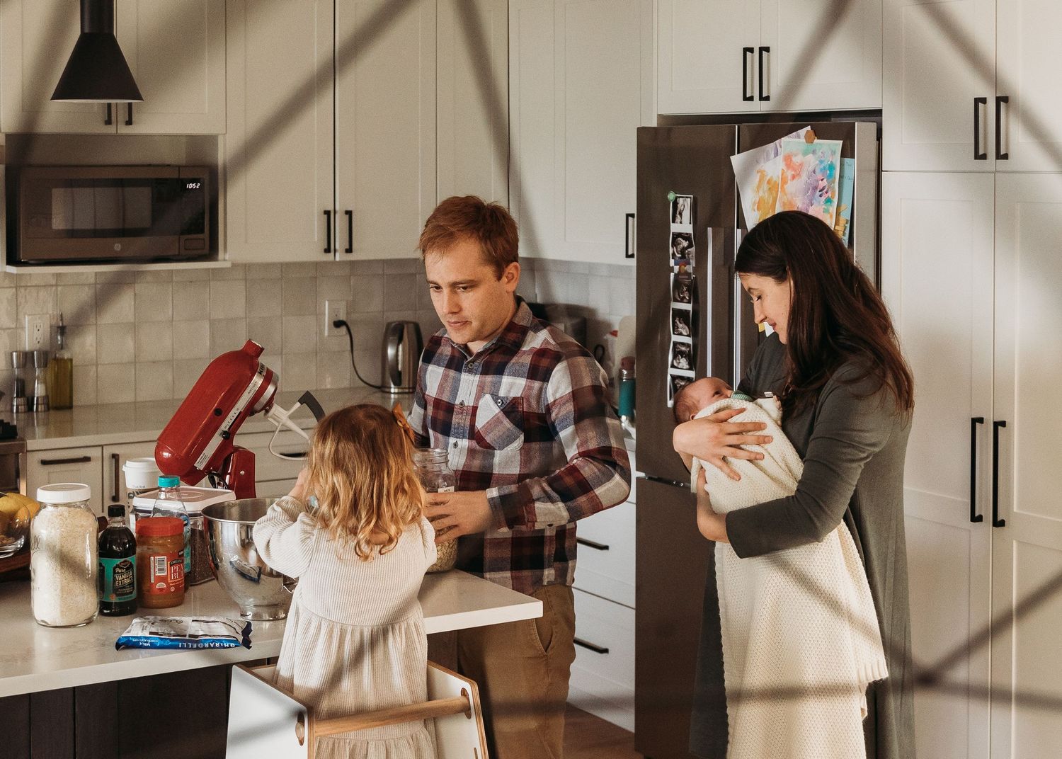 Family in modern kitchen, parents with newborn baby and young daughter baking together at counter.
