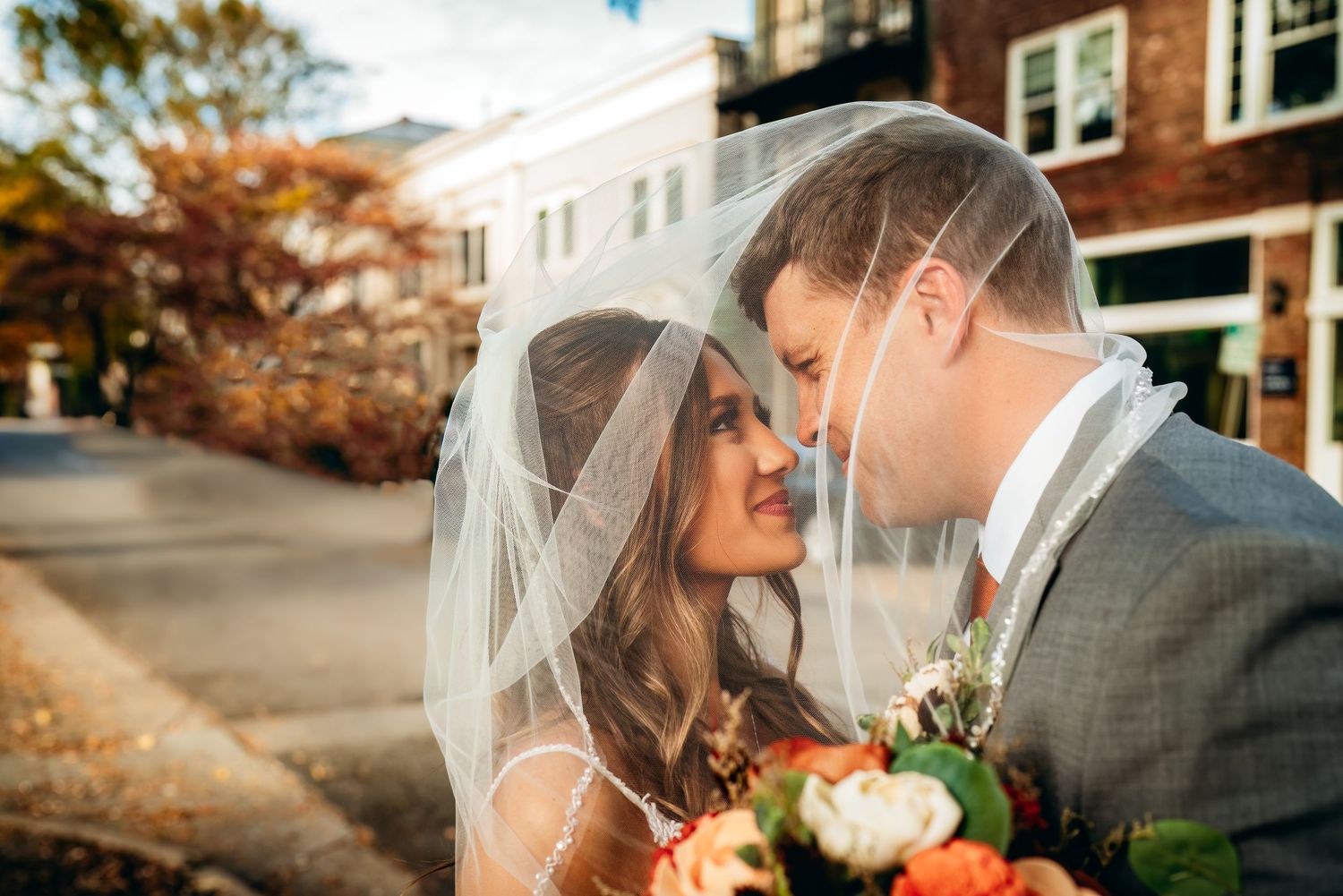 Bride and groom share a romantic moment under a wedding veil outdoors in autumn, with colorful fall foliage in the background.