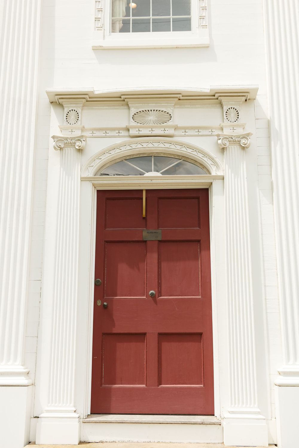 Red double door with ornate white colonial architectural surround, pilasters, and fanlight window on white building.