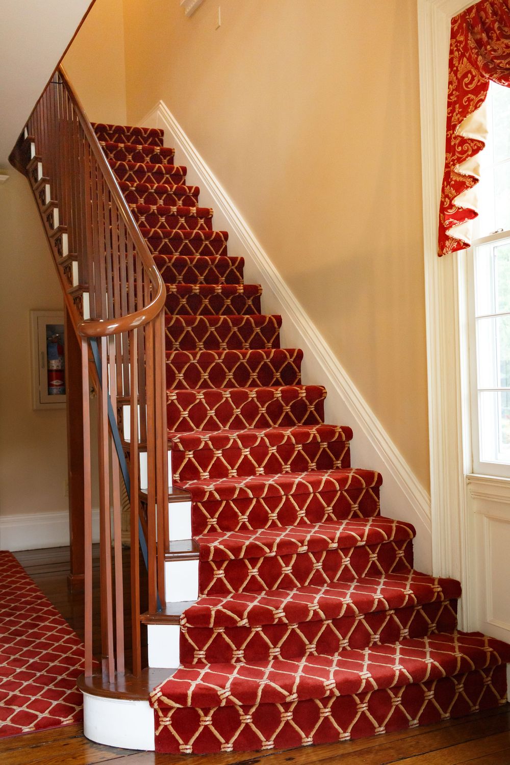 Elegant staircase with red and gold patterned carpet runner, wooden banister, and white risers in a classic home interior.