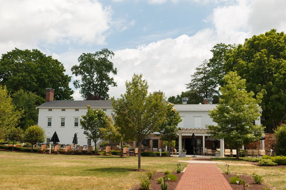 White colonial estate with brick pathway, lush green trees, and outdoor seating area on a sunny summer day.