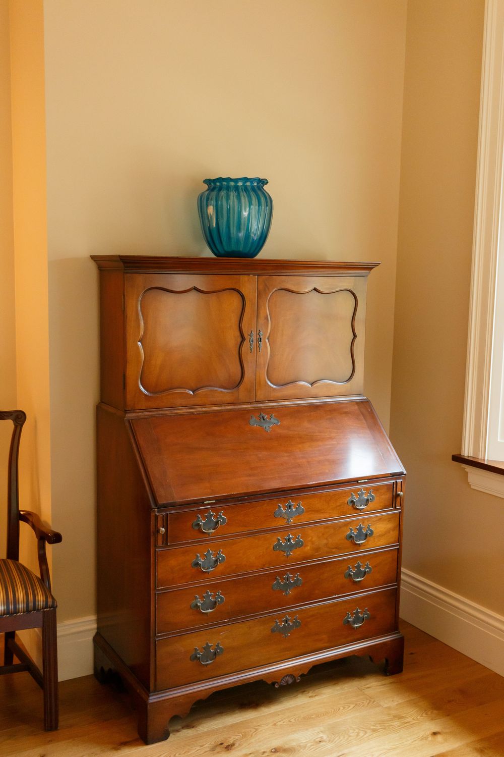 Antique wooden secretary desk with brass hardware and teal vase, set in a warm-toned corner room with hardwood floors.