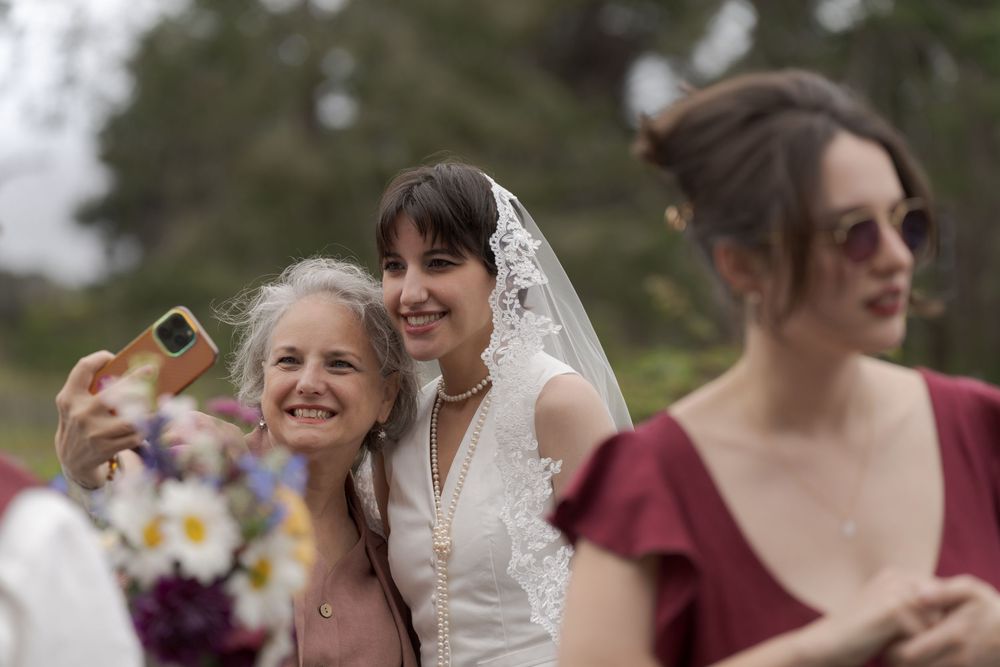 Bride in white dress and veil takes selfie with guest at outdoor wedding while bridesmaid in burgundy stands nearby.