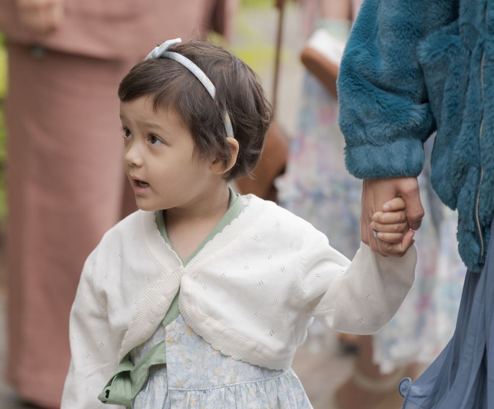 Young girl in white cardigan and headband holds an adult's hand at an outdoor gathering.