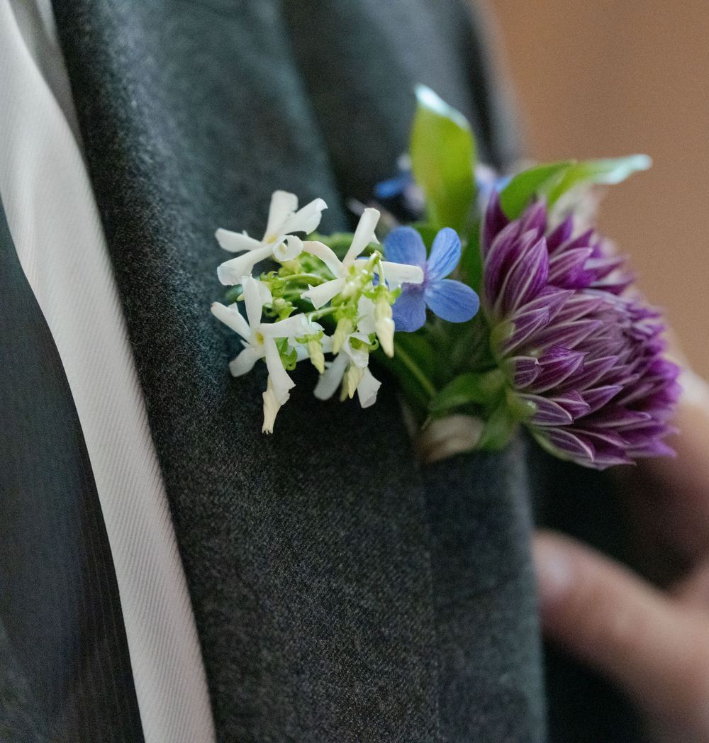 Close-up of a wedding boutonniere featuring white, blue, and purple flowers pinned to a gray suit lapel.