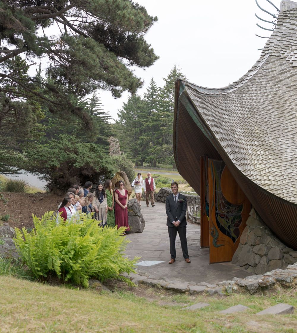 Wedding guests gather outside a unique curved roof chapel surrounded by pine trees for an outdoor ceremony.