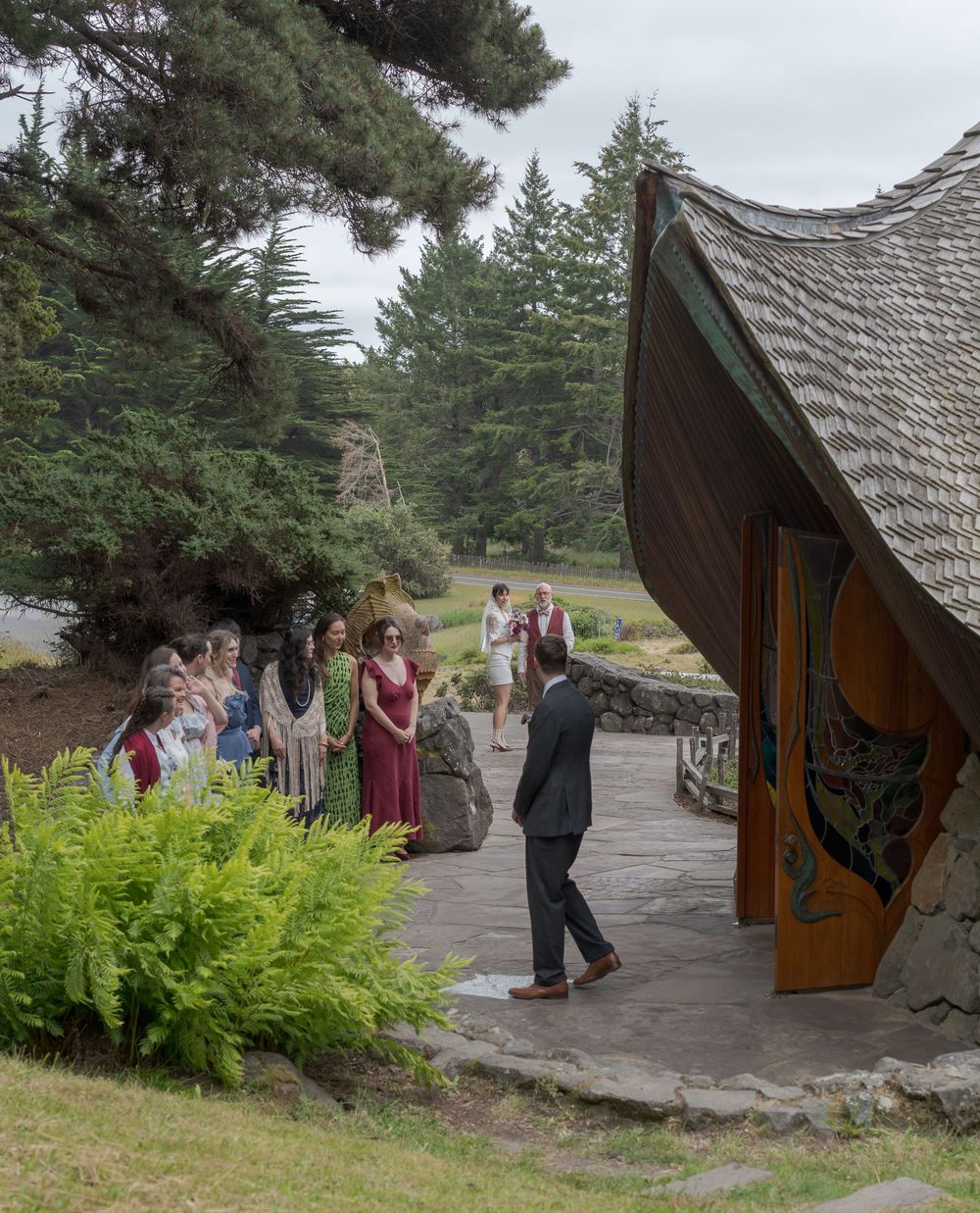 Outdoor wedding ceremony at a rustic chapel with guests gathered on stone patio surrounded by evergreen trees.