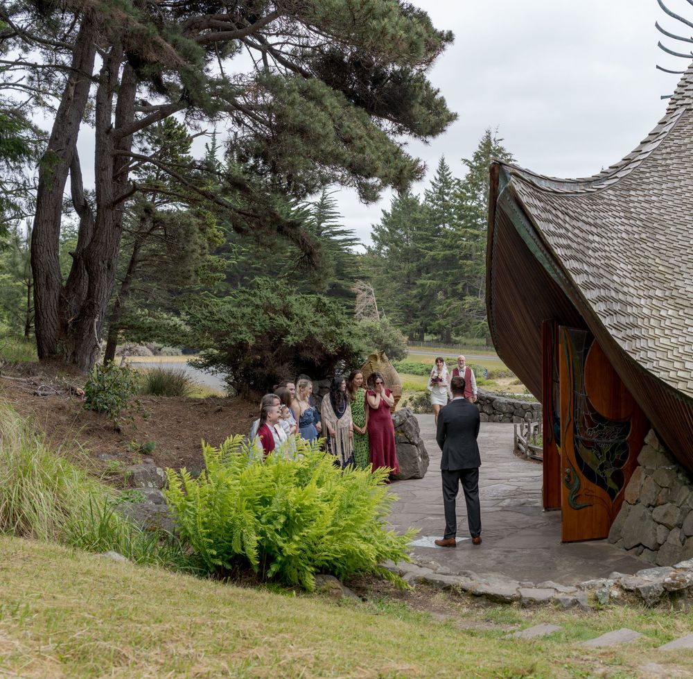 Wedding party gathered outside a unique thatched-roof venue surrounded by lush pine trees and ferns on a cloudy day.