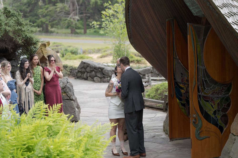 Couple sharing a romantic moment at an outdoor wedding ceremony near a rustic wooden chapel surrounded by lush greenery.
