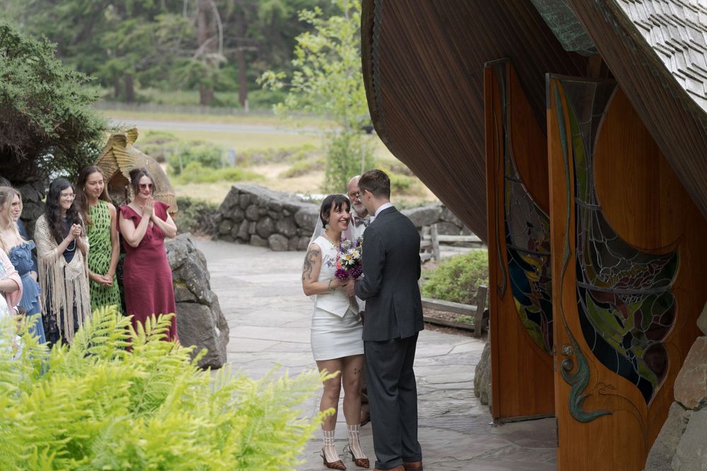 Couple exchanges vows at outdoor wedding ceremony near ornate wooden doors with guests and musician in background.