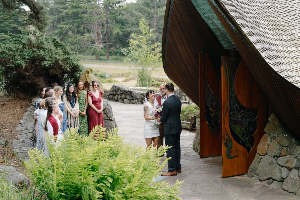 Small outdoor wedding ceremony with guests gathered near a rustic wooden chapel surrounded by lush greenery.