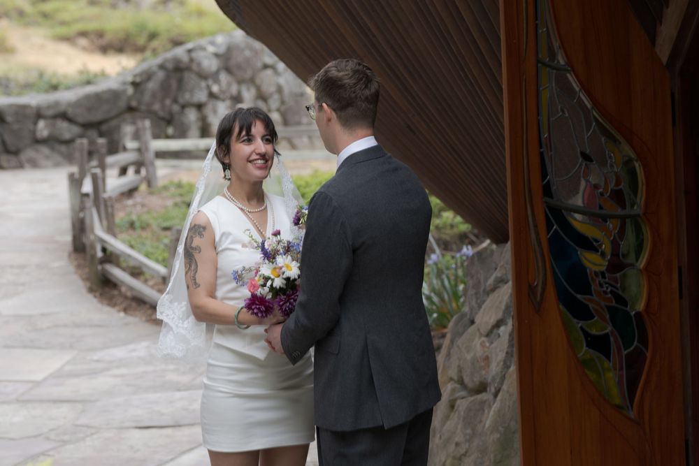 Bride in white dress holding bouquet smiles at groom in gray suit during outdoor wedding ceremony.
