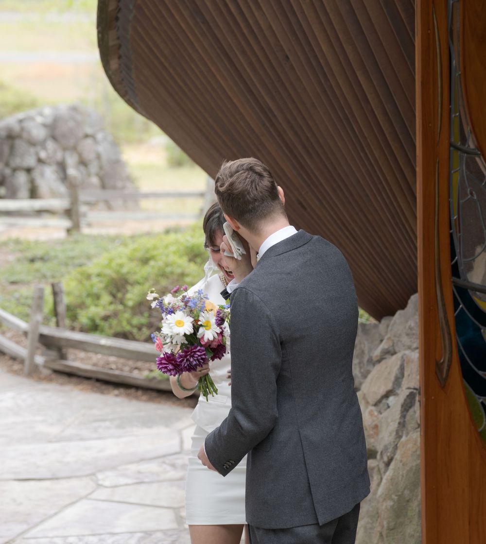 A bride holding a colorful bouquet and groom in gray suit share an intimate moment outside a rustic wooden venue.