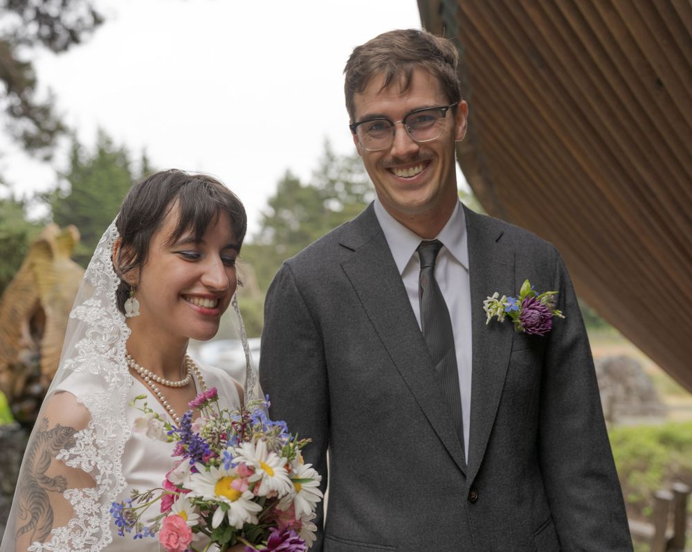 Smiling bride and groom pose outdoors on their wedding day, bride holding colorful bouquet.