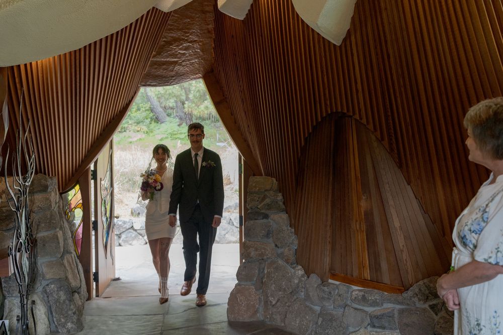 Bride and groom walking into a rustic wood-paneled wedding venue with stone walls and natural light.