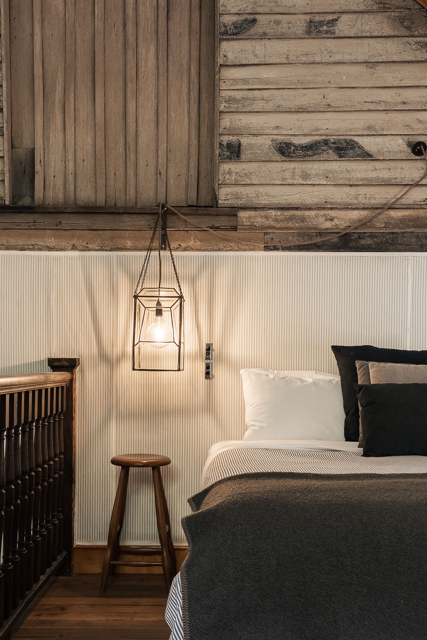 Cozy rustic bedroom with industrial cage pendant light, wooden walls, and dark gray bedding.

The Barn