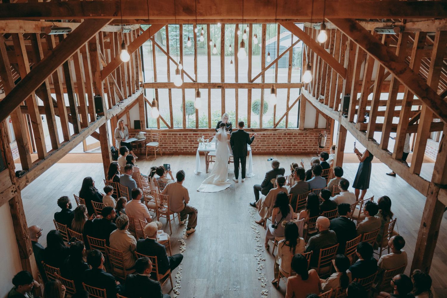 Aerial view of a rustic barn wedding ceremony with guests seated in rows beneath wooden beams.