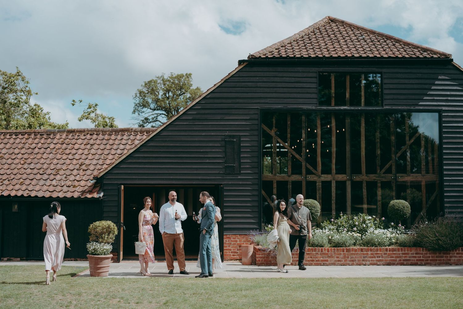 Guests mingling outside a modern black barn wedding venue with terracotta roof and string lights.