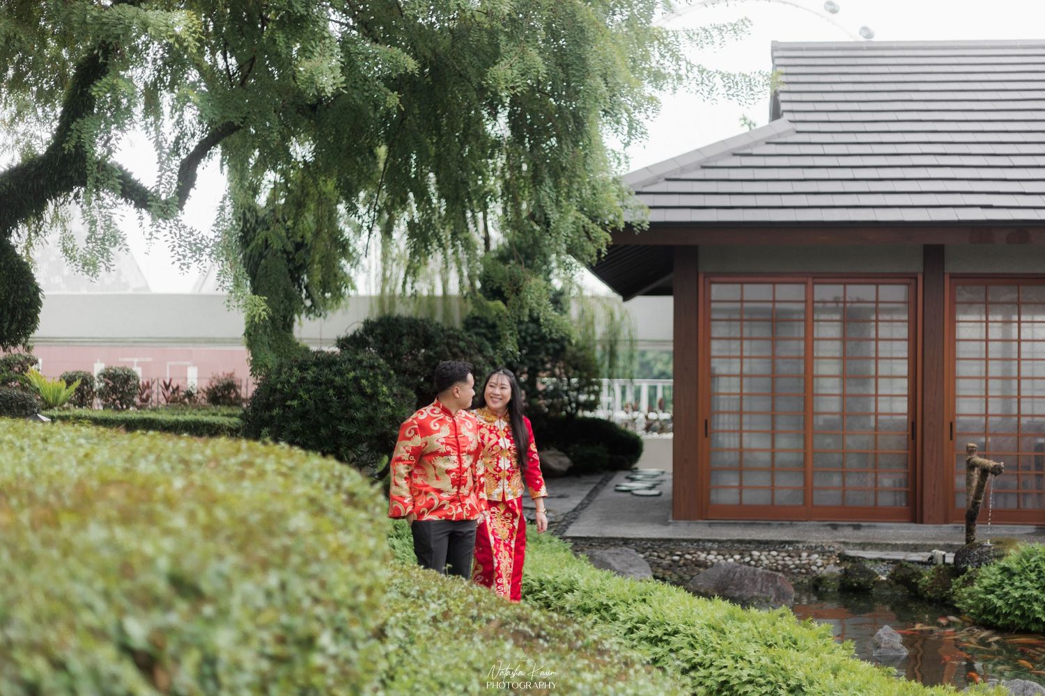 Couple in traditional red Chinese wedding attire walking through a lush Japanese garden near a wooden building.