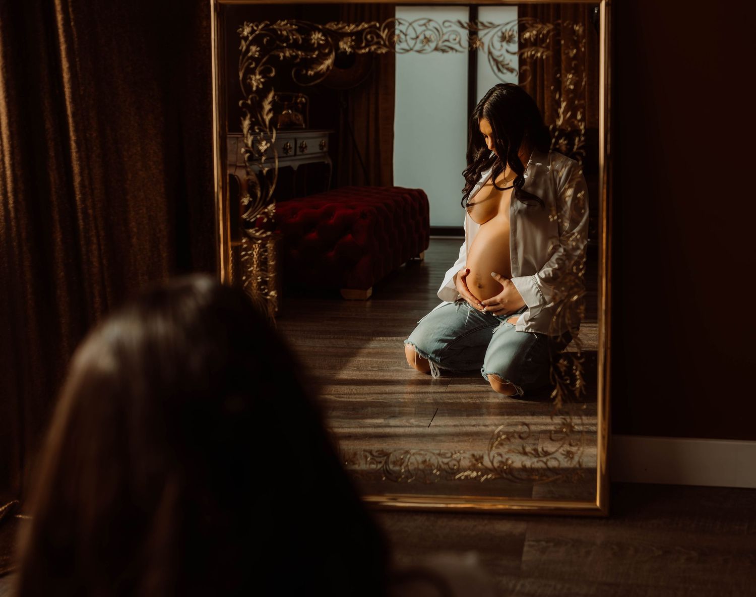 Pregnant woman admiring her baby bump reflection in an ornate gold-framed mirror in a warmly lit room with Boudoir by KJ.